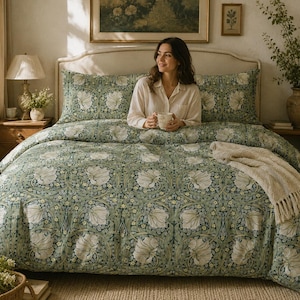Op de afbeelding: Een bed met een groen en wit bloemmotief dekbedovertrek en kussenslopen. Een crèmekleurige deken is over het bed gedrapeerd. De kamer is ingericht met een lamp, kunstwerken en een vaas met groen.