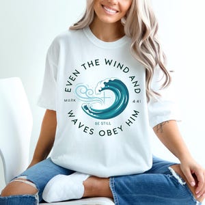 May include: White t-shirt with a teal and blue wave design and the text "Even the wind and waves obey him" in a circular layout. The shirt also features the text "Mark 4:41" and "Be Still".