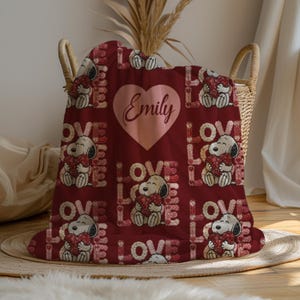 May include: A burgundy blanket featuring a repeating pattern of Snoopy holding a heart, the word "LOVE", and a large pink heart with the name "Emily". The blanket is folded in a wicker basket.