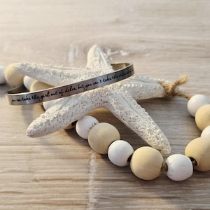 May include: A silver bracelet with the inscription "you can take the girl out of delco, but you can't take the delco out of the girl" rests on a white starfish. A strand of wooden beads in white and tan tones is also present.