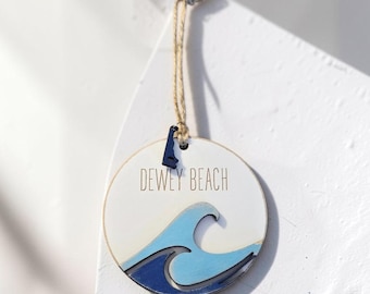 Puede incluir: Un adorno redondo de madera blanca con el texto DEWEY BEACH en letras negras. El diseño incluye formas de olas recortadas y superpuestas en azul claro y azul oscuro, con un pequeño dije negro con forma de estado unido al cordel de yute.