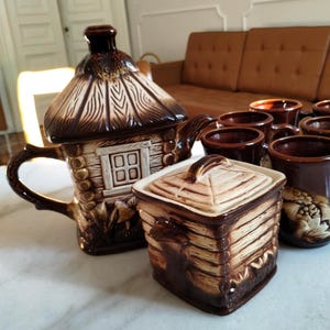 Op de afbeelding: Een bruine keramische theeset met een hutontwerp. De set bevat een theepot, een suikerpot en verschillende mokken. De theepot heeft een rieten dak en een klein raam. De mokken hebben een decoratief reliëf ontwerp.