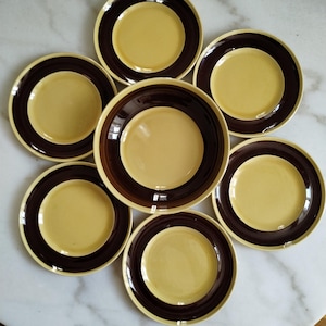 May include: A set of seven round ceramic plates arranged in a circular pattern. Each plate features a smooth, pale yellow center surrounded by a wide, dark brown rim. The plates have a glossy finish and a simple, uniform design across all pieces.