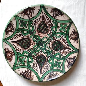 Peut inclure: Une assiette ronde en céramique avec un motif symétrique détaillé. L'assiette présente un fond crème, une bordure verte et des motifs complexes verts et bruns. Le motif comprend des éléments floraux et géométriques.
