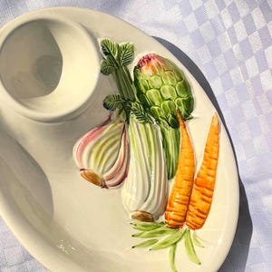 Può includere: Piatto da portata in ceramica color crema con una ciotola per salse integrata. Il piatto è decorato con immagini a rilievo di verdure, tra cui carote, carciofi, sedano e aglio. Le verdure sono dipinte con colori naturali.
