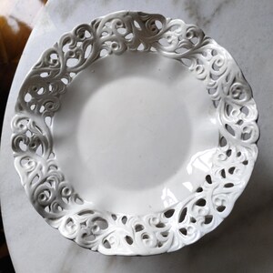 Op de afbeelding: Een witte keramische plaat met een decoratieve, opengewerkte rand. De rand heeft een herhalend bloemen- en scrollontwerp. De plaat heeft een gladde, glanzende afwerking en is geplaatst op een marmeren oppervlak. Deze plaat is geschikt voor het serveren van voedsel of als decoratief item.