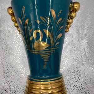 Puede incluir: Jarrón de color turquesa con detalles dorados. Presenta un cisne dorado y juncos. Tiene asas doradas con cuentas a cada lado. La base y el borde también son dorados. El jarrón mide unos 20 cm de alto.