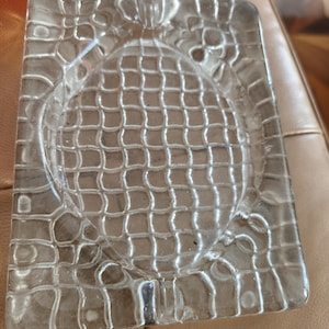 Op de afbeelding: Transparante glazen asbak met een rechthoekige vorm en een cirkelvormige inkeping in het midden. De asbak heeft een gestructureerd, rasterachtig patroon en een decoratieve rand.