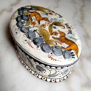 Może przedstawiać: Owalne ceramiczne pudełko o kremowej podstawie z ręcznie malowanymi ilustracjami dwóch pomarańczowych psów, szarymi okrągłymi motywami i złotymi akcentami kwiatowymi. Boki ozdobione są czarnymi, szarymi wzorami oraz pomarańczowymi kropkami.