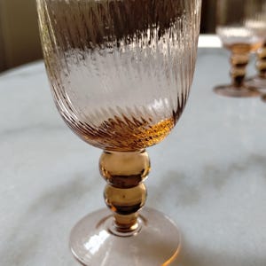 Può includere: Un set di quattro calici ambrati. Ogni bicchiere presenta un design strutturato e a coste sulla coppa e uno stelo composto da tre sfere impilate. La base è di forma piatta e circolare. I bicchieri sono disposti su una superficie bianca.