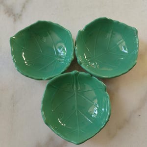 Può includere: Tre ciotole in ceramica verde a forma di foglia. Ogni ciotola ha una finitura lucida e dettagliati motivi di venature delle foglie. Le ciotole sono disposte in una formazione triangolare, adatte per servire piccole porzioni di cibo o come elementi decorativi.