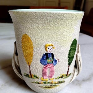 Peut inclure: Un mug en céramique blanc cassé à la texture granuleuse, orné d'une illustration peinte représentant une personne aux cheveux blonds vêtue d'une chemise à carreaux bleus, d'un gilet sombre et d'un pantalon à carreaux roses, entre deux arbres.