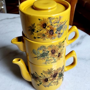 Könnte beinhalten: Ein Stapel aus drei gelben Keramikgefäßen mit einem aufgemalten Sonnenblumenmuster. Das oberste Teil ist eine Schüssel mit Deckel, während das mittlere und das untere Teil Krüge mit Ausgüssen und Griffen sind. Jedes Teil zeigt gelbe Blumen.