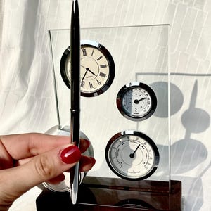 Könnte beinhalten: Eine Tischuhr mit Quarzuhr, Thermometer und Hygrometer. Die Uhr hat ein weißes Zifferblatt mit schwarzen römischen Ziffern. Thermometer und Hygrometer haben schwarze Zifferblätter mit weißen Markierungen. Ein silberner Stift wird vor der Uhr gehalten.