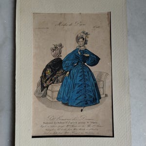 Könnte beinhalten: Ein antiker Mode-Druck mit dem Titel "Modes de Paris", der zwei Damen in eleganter Kleidung zeigt. Eine Dame trägt ein leuchtend blaues Kleid mit passendem Hut, die andere ein schwarzes Ensemble. Der Druck ist in einem neutral getönten Passepartout gerahmt.