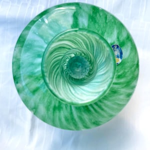Puede incluir: Vista superior de un jarrón de vidrio verde con un patrón de remolino. El jarrón tiene una base ancha y plana y un cuerpo redondeado que se estrecha hacia la parte superior. El vidrio es una mezcla de verde claro y oscuro, con remolinos blancos.