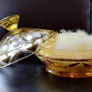 Könnte beinhalten: Ein Vintage-Puderbehälter aus bernsteinfarbenem Glas mit passendem Deckel und Quaste. Die Dose und der Deckel haben geätzte Blumen- und geometrische Designs. Die Quaste ist hellgelb und flauschig.