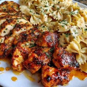 Pode incluir: Um prato de comida com frango grelhado e massa. O frango é cortado em pedaços e tem uma cobertura marrom avermelhada. A massa tem formato de laço e é coberta com um molho cremoso, com ervas verdes polvilhadas por cima. A comida está em um prato branco.