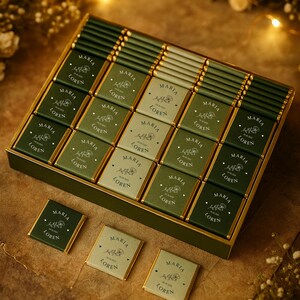 Könnte beinhalten: Eine Schachtel Pralinen mit einzeln verpackten Quadraten in Grün und Gold. Jedes Schokoladenquadrat ist mit den Namen "MARIA" und "LOREN" in weißer Schrift sowie einem floralen Design versehen. Die Schachtel ist grün mit einem goldenen Rand.