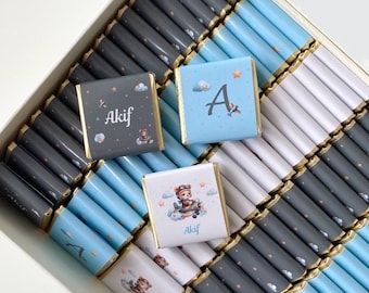 May include: A box of chocolates with blue, white, and gray wrappers. Some chocolates have the name "Akif" printed on them, while others have the letter "A" on them. The chocolates are arranged in rows.