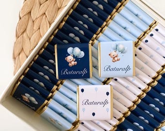 May include: A white box filled with individually wrapped chocolates. The chocolates are in shades of blue and white, some with a teddy bear and balloon design, and the name "Baturalp". The box sits on a woven wicker surface.