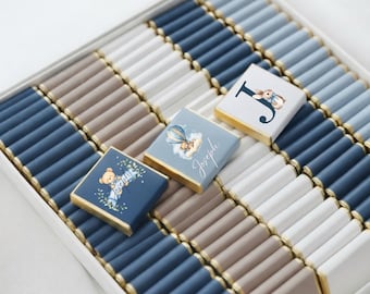 May include: A box of chocolates with a variety of colors, including blue, white, and brown. Some chocolates have decorative designs, including a teddy bear, a hot air balloon, and a letter "J" with a bunny. The chocolates are arranged in rows.