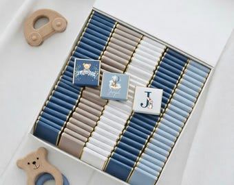 May include: A white box filled with rows of blue, gray, and white chocolate bars. Three decorated chocolates are visible, one with a teddy bear, one with a hot air balloon, and one with the letter "J" and a bunny. Wooden baby toys are also present.