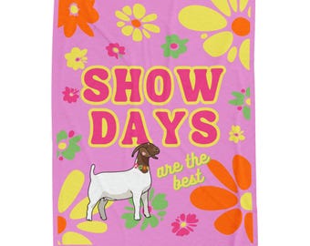 Puede incluir: Una manta rosa con las palabras "SHOW DAYS" en amarillo y rosa, y una ilustración de una cabra. La manta tiene un diseño floral en naranja, amarillo y verde. También se incluye el texto "are the best".