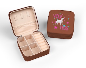 Puede incluir: Una caja de joyería marrón, abierta para revelar compartimentos y porta anillos, se encuentra junto a una caja cerrada. La caja cerrada presenta una ilustración de perro blanco con el texto "YOU" y "THIS" en rosa y azul, rodeado de estrellas.