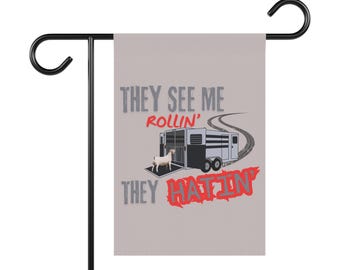 Puede incluir: Una bandera de jardín gris con un soporte de metal negro. La bandera presenta el texto "THEY SEE ME ROLLIN' THEY HATIN'" con una ilustración de una cabra, un remolque y huellas de neumáticos.