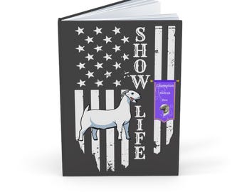 Puede incluir: Un cuaderno con una cubierta gris oscuro que presenta una ilustración de cabra blanca y las palabras "SHOW LIFE" en blanco. El diseño de fondo se asemeja a la bandera estadounidense. Una pequeña pancarta púrpura dice "Champion Overall Doe".
