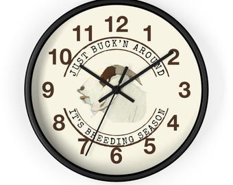 Puede incluir: Reloj de pared redondo con marco negro y esfera color crema. El reloj presenta una ilustración de cabra y el texto "JUST BUCK'N AROUND" y "IT'S BREEDING SEASON". Las manecillas del reloj son negras.