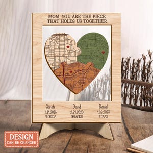 Könnte beinhalten: Holzgerahmtes Herz-Kartenkunstwerk mit dem Text "MOM, YOU ARE THE PIECE THAT HOLDS US TOGETHER". Das Herz ist in drei Abschnitte mit unterschiedlichen Kartendesigns unterteilt. Darunter Namen, Daten und Orte. Das Design kann geändert werden.