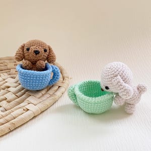 May include: Crocheted toys featuring a brown dog in a blue teacup and a light gray dog interacting with a green teacup. The toys are handmade and sit on a woven mat, with a neutral background.