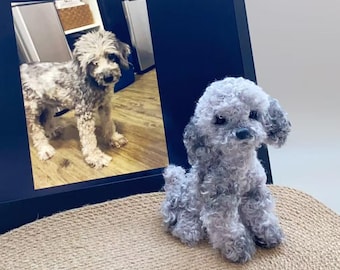 May include: A grey, plush toy poodle sits on a woven mat beside a laptop displaying a photo of a similar dog. The toy has curly fur and dark eyes. The laptop screen shows a photo of a dog with a similar coat pattern.