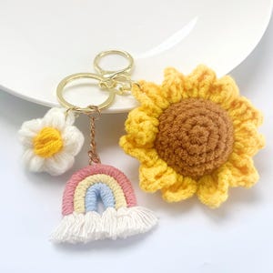 May include: A handmade keychain featuring a crochet sunflower, daisy, and rainbow charm. The sunflower has yellow petals and a brown center. The daisy is white with a yellow center. The rainbow is pastel-colored with white fringe. The keychain has a gold-colored ring.
