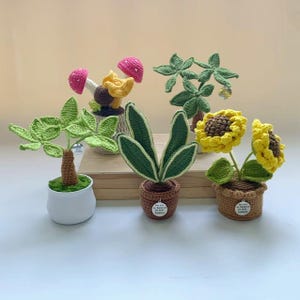 May include: A collection of handmade crochet plants, including a sunflower, snake plant, and mushroom. The plants are in small pots and feature vibrant colors like yellow, green, and pink. The crochet details are intricate and add a unique touch.