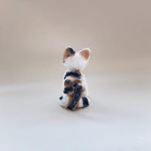 以下が含まれることがあります： 後ろから見た手作りのフェルト猫の置物。猫は白で、茶色、黒、タンの模様があります。尖った耳と長い白いひげがあります。猫は中立的な表面に直立して座っています。