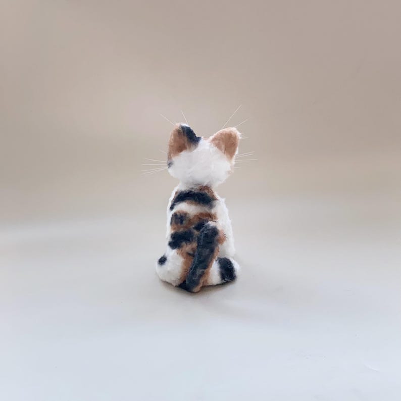 以下が含まれることがあります： 後ろから見た手作りのフェルト猫の置物。猫は白で、茶色、黒、タンの模様があります。尖った耳と長い白いひげがあります。猫は中立的な表面に直立して座っています。