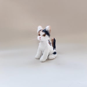 以下が含まれることがあります： 白、茶色、ネイビーブルーの模様が入った、座っている猫のぬいぐるみ。猫は長い白いひげ、黄色の目、ピンクの鼻をしています。耳と尾には茶色とネイビーブルーのパッチがあります。