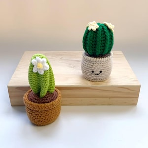 May include: Two crocheted cactus plants in pots. One is green with a white flower and a brown pot. The other is a green cactus with white flowers in a beige pot with a smiling face. The plants are on a wooden surface.