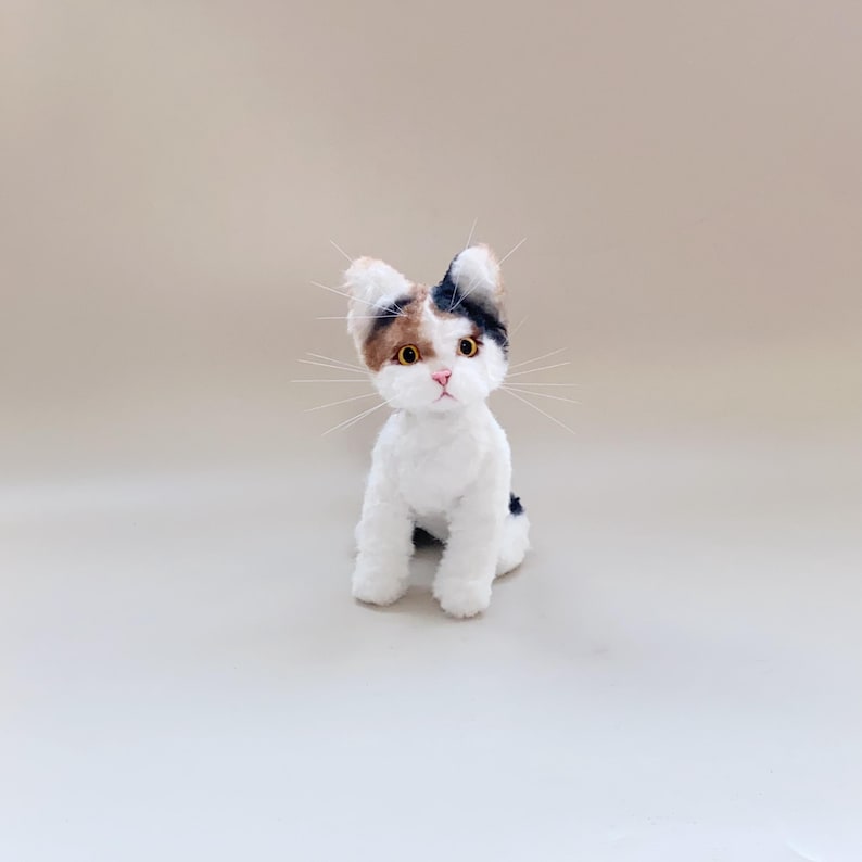 以下が含まれることがあります： 手作りのぬいぐるみキャリコ猫の置物。猫は白く、頭と背中に茶色と黒の毛皮のパッチがあります。黄色い目と長い白いひげがあります。猫は直立しています。