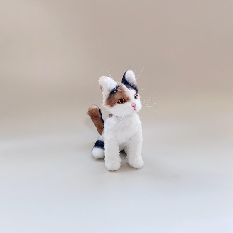 以下が含まれることがあります： 手作りのぬいぐるみキャリコ猫の置物。猫は白で、茶色、黒、タンの毛皮のパッチがあります。黄色い目、ピンクの鼻、長い白いひげがあります。猫は尻尾を丸めて直立しています。