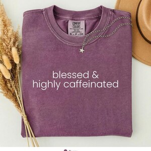 Op de afbeelding: Een bessenkleurig t-shirt met korte mouwen en een ronde hals, voorzien van de witte tekst 'blessed & highly caffeinated' in het midden op de borst. Een dunne zilverkleurige ketting met een kleine sterhanger rust op de kraag.