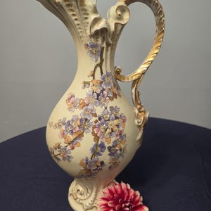 May include: A cream-colored ceramic pitcher featuring a decorative gold-trimmed handle and a scalloped rim. The body is adorned with painted floral clusters in shades of purple, pink, and orange, accented with gold detailing throughout.
