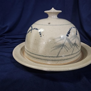 May include: A ceramic cloche with a matching plate. The cloche is off-white with blue and gray painted accents and a small knob on top. The plate is round and the same off-white color. The cloche and plate are sitting on a dark blue surface.