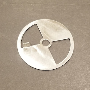 May include: A circular, silver-colored metal object with a cut-out design. The design features a central hole and three pie-shaped sections, with a small rectangular notch on the edge. The object has a brushed metal finish.