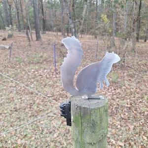 May include: A metal squirrel silhouette mounted on a wooden post. The squirrel is facing to the right and has a bushy tail. The post is surrounded by brown leaves and a wire fence.
