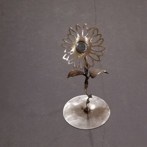 May include: A metal sunflower sculpture with a circular base. The sunflower is made of thin metal and has a detailed design. The base is a flat circle with a slightly raised center.
