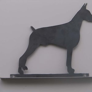 May include: A dark gray metal silhouette of a Doberman Pinscher dog. The dog is depicted in a standing pose, with its tail slightly raised. The silhouette is mounted on a rectangular base.